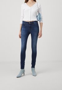 Vaqueros ajustados de color azul oscuro combinados con una blusa blanca de manga larga y con canalé, que presenta un escote en V profundo, y botas de tobillo puntiagudas de color azul claro.