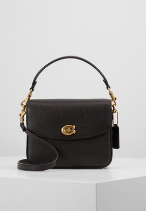 Sac à main en cuir noir avec fermoir doré, poignée unique sur le dessus, bandoulière amovible et petite étiquette suspendue, présenté sur un piédestal blanc.