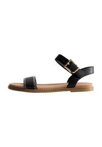 Sandalias de cuero negro con suela plana, que cuentan con una amplia correa frontal, una correa para el tobillo con una hebilla dorada y un detalle de costura blanca.