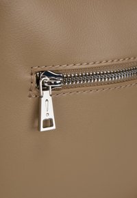 Beige imitation læderpose med en sølvfarvet lynlås med en karakteristisk trækflap og metalstænger, syet detalje ved lynlåsen.