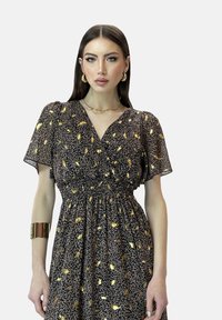 Robe noire avec un motif floral doré, manches courtes à volants, taille smockée, tissu léger, portée avec un bracelet doré et des colliers superposés.