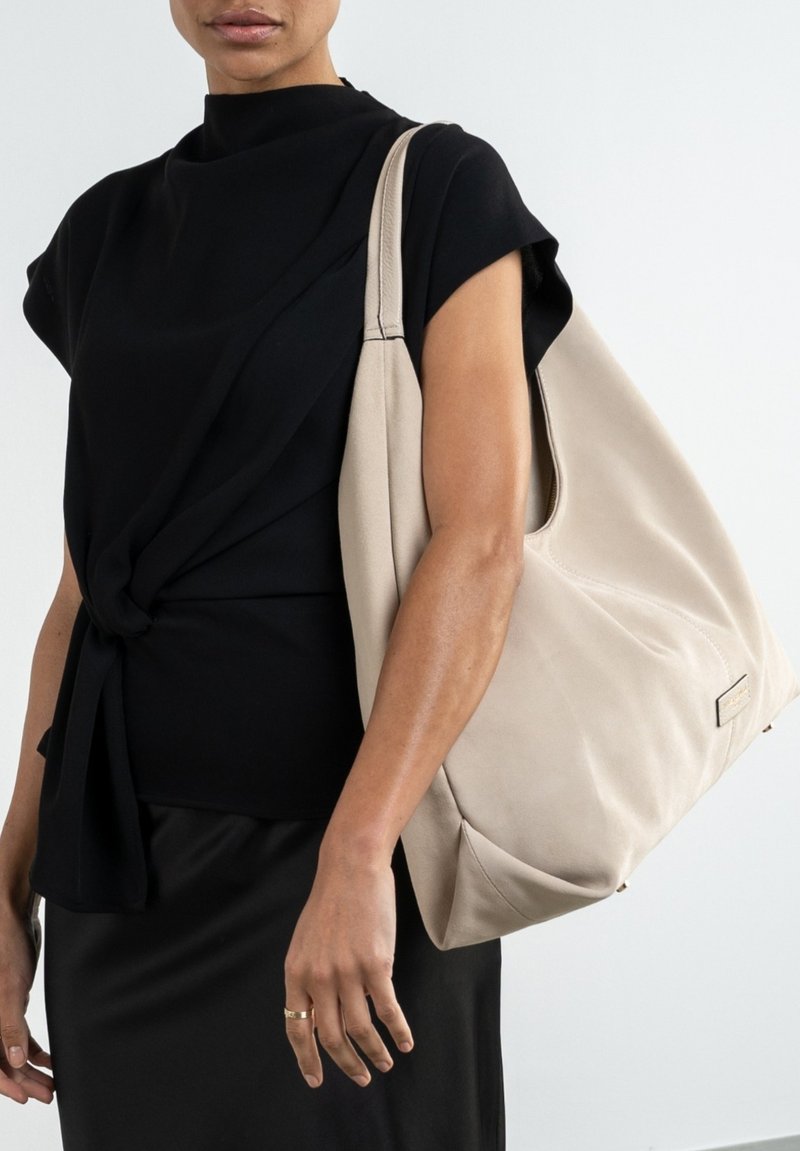 Beige, lässige Handtasche aus weichem Material mit runder Form, großem Öffnung und seitlichem Logo-Detail. Trägt man über einem schwarzen Oberteil.