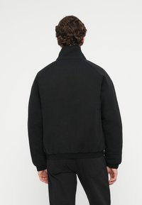 Black bomber jacket with a high collar, textured fabric, raglan sleeves, and elastic cuffs at the hem. Machine-washable for easy care.