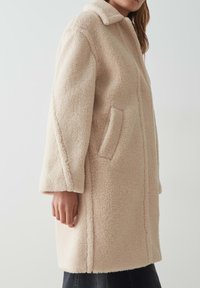 Cappotto beige chiaro realizzato in un materiale testurizzato e soffice. Presenta un colletto classico, tasche laterali e un taglio dritto e moderno.