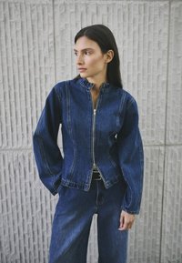 Jeans jakna s patentnim zatvaračem sprijeda, okomitim ovratnikom i dugim rukavima. Karakterizira je tamnoplava boja i vidljivo šivanje duž šavova.
