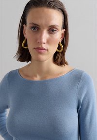 Vaaleansininen ribattu neulepaita, jossa on veneenkaulus, yhdistettynä suuriin, pyöreisiin kultaisiin korvaketjuihin. Sujuva materiaali ja istuva muotoilu.