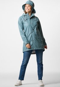 Vattentät teal regnjacka med huva, utrustad med knappstängningar, två sidofickor och en längre bakre fåll. Matchad med avslappnade blå jeans.