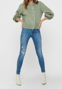 Zielony sweter z dzianiny z oversize'owymi rękawami i sześcioma okrągłymi guzikami, zestawiony z niebieskimi, przetartymi rurkami i białymi botkami za kostkę.