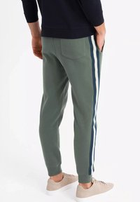 Grüne Jogginghosen mit seitlichen Streifen in Marineblau und Weiß. Hergestellt aus weichem Stoff mit schmaler Passform, gerippten Bündchen und einer Detailtasche auf der Rückseite.