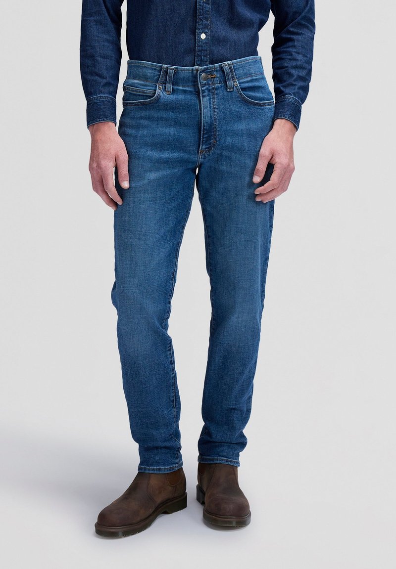 Homme portant un jean en denim bleu, des bottes marron foncé et une chemise en denim bleu foncé, debout contre un fond clair uni.