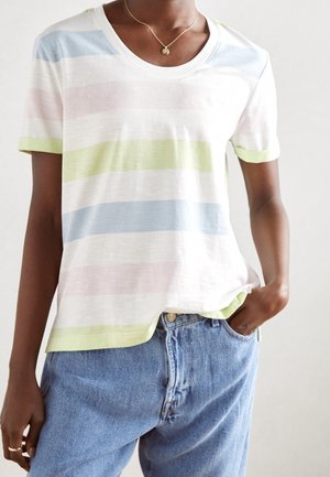 Personne portant une chemise à manches courtes rayée aux tons pastel, un jean bleu, un collier pendentif en or et des bagues, une main dans la poche, sur un fond uni.