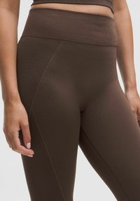 Leggings bruns avec une taille haute côtelée, présentant un motif texturé sur les cuisses et une finition lisse, conçus pour le confort et la flexibilité.