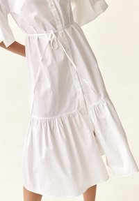 Abito bianco con bottoni e maniche corte, dotato di vita legata, gonna arricciata e orlo asimmetrico. Realizzato in tessuto leggero e morbido.
