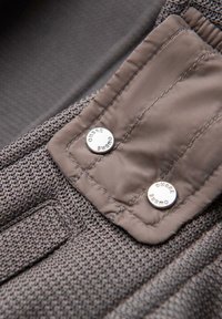 Gros plan sur un tissu tricoté marron avec un rabat en tissu cousu, fixé par deux boutons argentés gravés du mot « OMERE ».