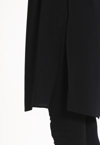 Svart tunika med avslappnad passform, försedd med en sidofålla, slät textur och längre baktill, designad för komfort och rörelsefrihet.