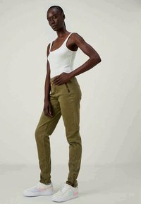 Mulher em pé, vestindo uma camisola branca sem mangas, calças verde-oliva com detalhes em zíper e sapatilhas brancas com acabamentos em tons pastel.