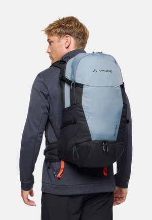 Homme portant un grand sac à dos Vaude bleu et noir avec des sangles de poitrine et de taille, vêtu de vêtements d'extérieur sombres, faisant face à gauche.
