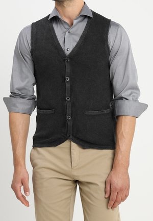 Mann trägt eine dunkelgraue, geknöpfte Strickweste über einem hellgrauen, hochgekrempelten Hemd mit beigen Hosen, steht vor einem einfarbigen Hintergrund.