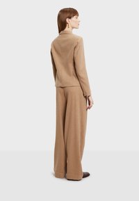 Béžový vlněný blazer s vysokým límcem a přiléhavým střihem, doplněný širokými béžovými vlněnými kalhotami. Hladká textura a minimalistický design.