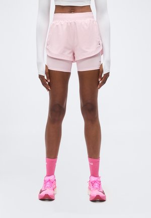 Persoon draagt lichtroze dubbele laag hardloopshorts, bijpassende roze sokken en roze sneakers, staand tegen een effen achtergrond.