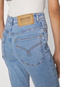 Jeans in denim azzurro con vita alta, tasche posteriori e cuciture arancioni a contrasto. Etichetta in pelle beige sulla vita con la scritta "Moschino Jeans".