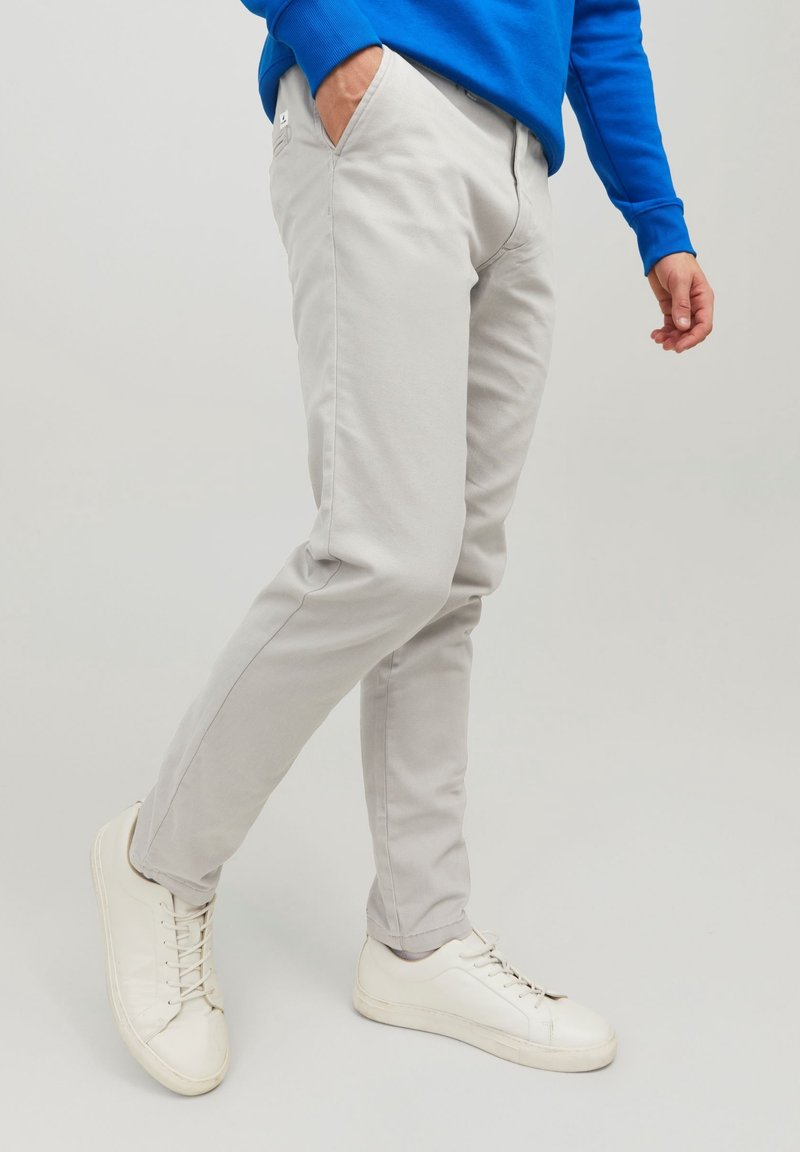 Personne portant un pantalon slim gris clair, des baskets blanches et un haut bleu à manches longues, debout avec une main dans la poche.