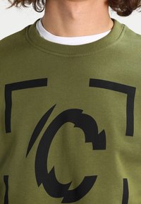 Olivově zelená mikina s kulatým výstřihem, s velkým černým grafickým potiskem uprostřed hrudi, vyrobená z měkkého bavlněného materiálu.