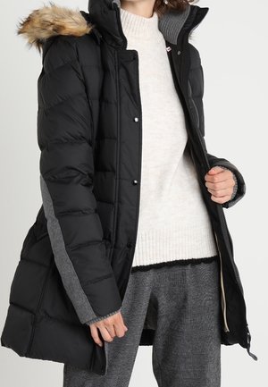 Kobieta w czarnej pikowanej kurtce z futrzanym kapturem, białym dzianinowym swetrze oraz szarych spodniach w kratę, zapinająca zamek kurtki jedną ręką.