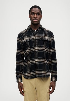 Mann in schwarz-beiger kariertem Hemd mit Druckknöpfen und beigen Cargo-Hosen, der vor einem einfarbigen, leichtem Hintergrund steht.