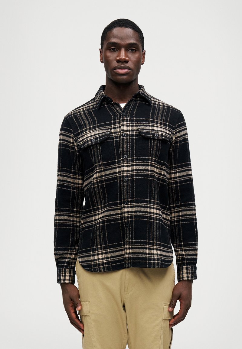 Hombre con una camisa de cuadros negra y beige de botones y pantalones cargo beige de pie contra un fondo claro liso.