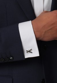 Navy suit jacket with a white cuff featuring a silver-toned cufflink in a crossed design. The fabric has a subtle grid pattern.