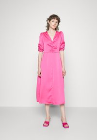 Vestido de cetim fúcsia com decote em V, mangas bufantes e detalhe de cintura franzida. Combinado com sandálias de slide transparentes em tom de rosa. Fundo minimalista.