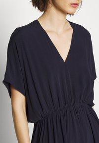 Marineblå kjole med dyb V-hals, løse ærmer og elastisk talje. Lavet af blødt stof med et subtilt struktureret mønster.