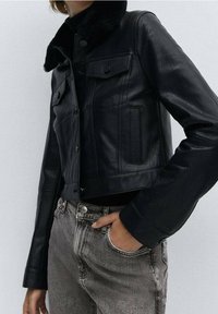 Chaqueta de cuero negra con un diseño recortado, cuello de piel, botones a presión y detalles de bolsillos, combinada con jeans de mezclilla grises en una pose casual.