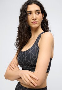 Γυναίκα με μακριά σγουρά μαλλιά, που φοράει μαύρο αθλητικό μπουστάκι με γεωμετρικά σχέδια, σταυρωμένα χέρια, κοιτάζοντας προς τα πλάγια με φόντο ένα απλό φόντο.