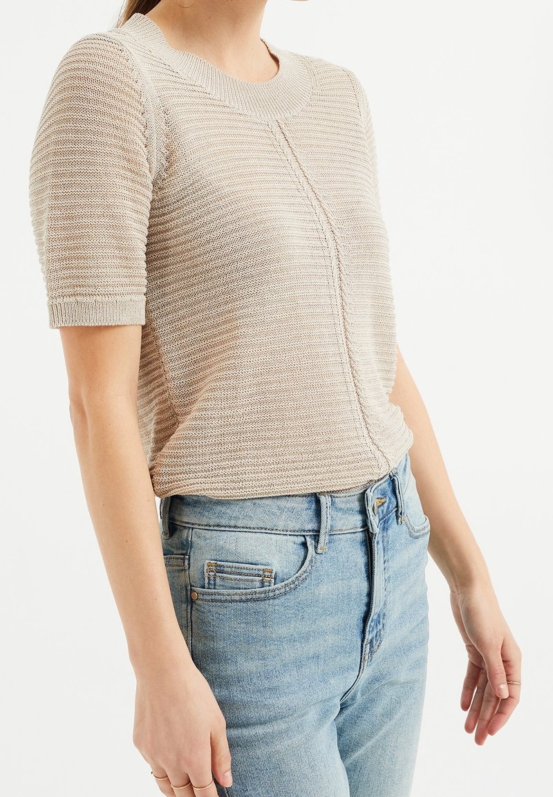 Beige stickad kortärmad topp med texturerat mönster, ribbad halsringning och rak passform, kombinerad med ljusblå midjehöga jeans.