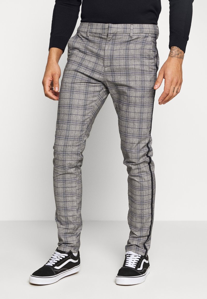 Homme portant un pantalon slim gris à carreaux avec une bande latérale noire et des baskets noires et blanches, debout devant un fond uni.