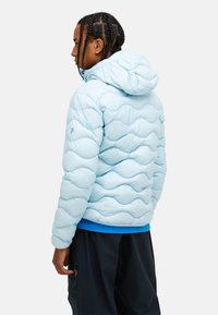 Lyseblå pufferjakke med et kviltet bølget mønster, justerbar hætte og elastiske manchetter. Båret over en blå skjorte og mørke bukser.