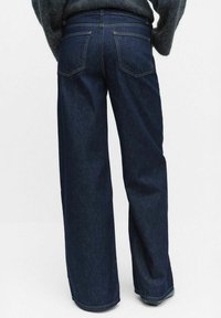 Vettore di jeans in denim scuro a gamba larga, con vita alta, doppie tasche posteriori e orlo pulito. Il tessuto ha una texture liscia con cuciture tono su tono.
