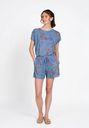 Vrouw draagt een blauwe wittebloemen korte mouw top en bijpassende korte broek met handen in de zakken, staand tegen een effen witte achtergrond.