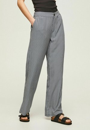 Femme portant un pantalon ample à motifs avec des poches et des sandales noires, se tenant devant un fond clair.