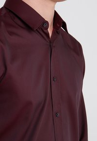 Camisa de botones burdeos con textura suave, cuello clásico y botones negros, que presenta un diseño ajustado y una apariencia nítida.