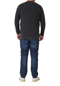 Mężczyzna ubrany w ciemnoszary sweter z dzianiny, niebieskie dżinsy i białe trampki, stojący tyłem na białym tle.