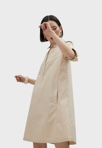 Abito camicia beige con colletto e maniche corte, dotato di chiusura frontale con bottoni e tasche laterali. Tessuto morbido con una silhouette comoda.