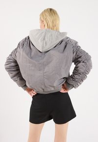 Graue, leichte Bomberjacke mit grauer Kapuze, gerafften Ärmeln und geripptem Saum. Getragen mit schwarzen Shorts, die einen lässigen Look zeigen.