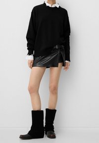 Suéter negro de gran tamaño con bordes deshilachados, superpuesto a una camisa blanca con cuello. Falda mini de cuero sintético negro con un corte asimétrico. Calentadores de piernas negros de punto.