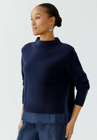 Dunkelblauer Pullover mit hohem Kragen, gerippten Bündchen und strukturiertem Finish. Über Jeans getragen, mit einem geschichteten Saumdetail.
