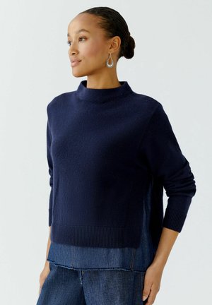 Navy blue sweater with a high neckline, ribbed cuffs, and a textured finish. Worn over denim, featuring a layered hem detail.