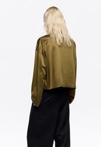Oliwkowa krótka bluzka z długim rękawem o gładkiej fakturze, luźnym kroju i szerokich rękawach, zestawiona z czarnymi spodniami o szerokich nogawkach.