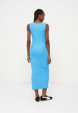 Kvinde med flettet hår iført en ærmeløs, lys blå midi-kjole og sorte og hvide flade sko, stående med ryggen mod os foran en hvid baggrund.